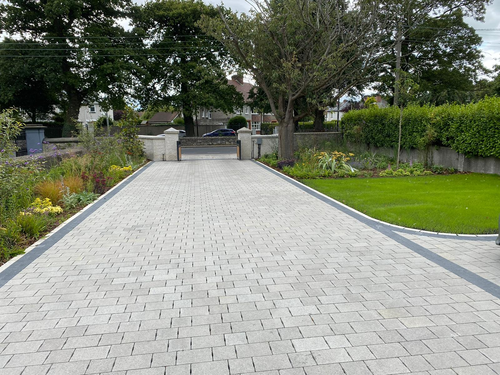 A block paved driveway running between clipped lawns and mature trees to a gated entrance