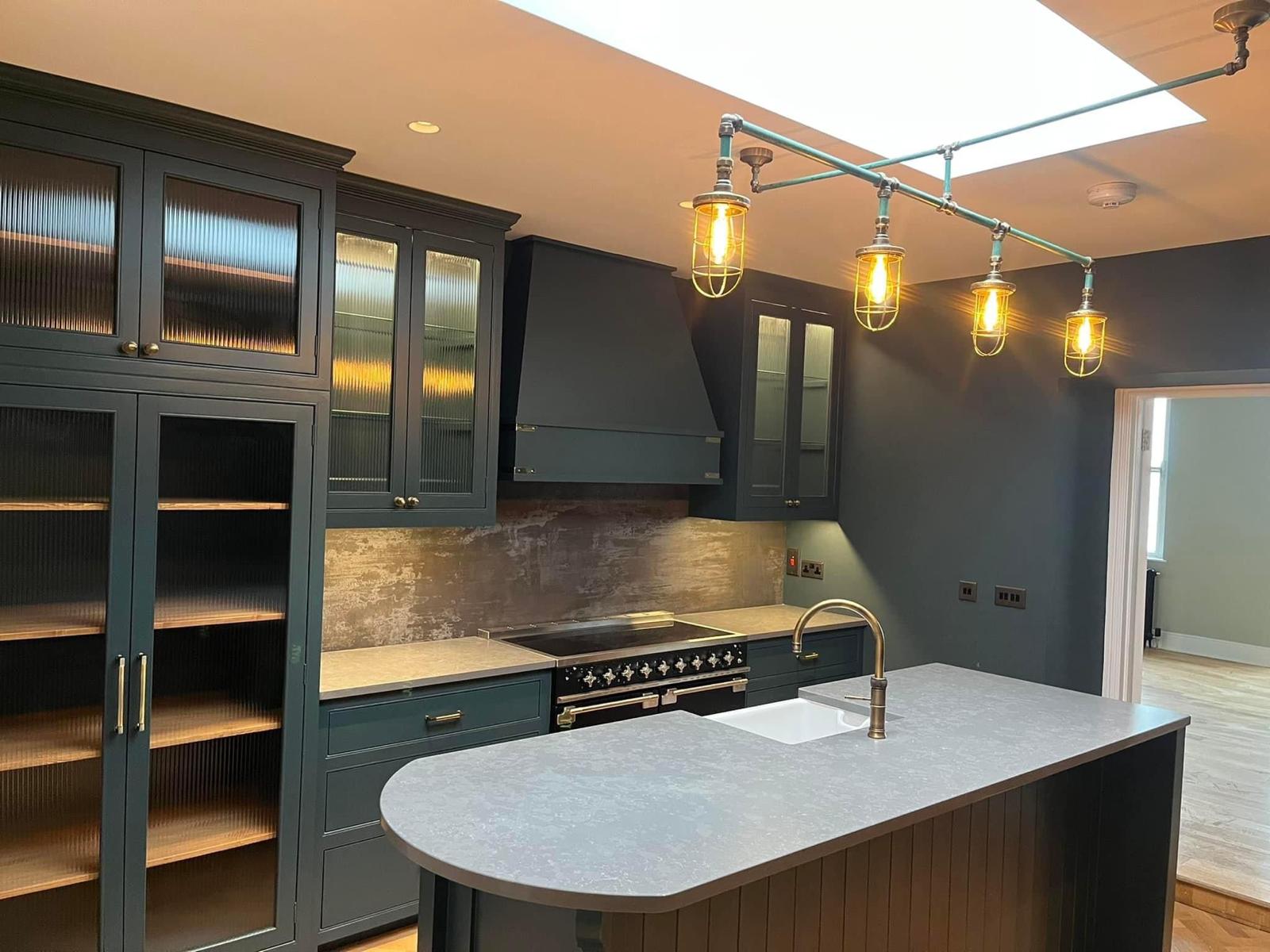 A kitchen with deep green cabinetry, brass tap, glazed wall units and pendant lights over a stone island