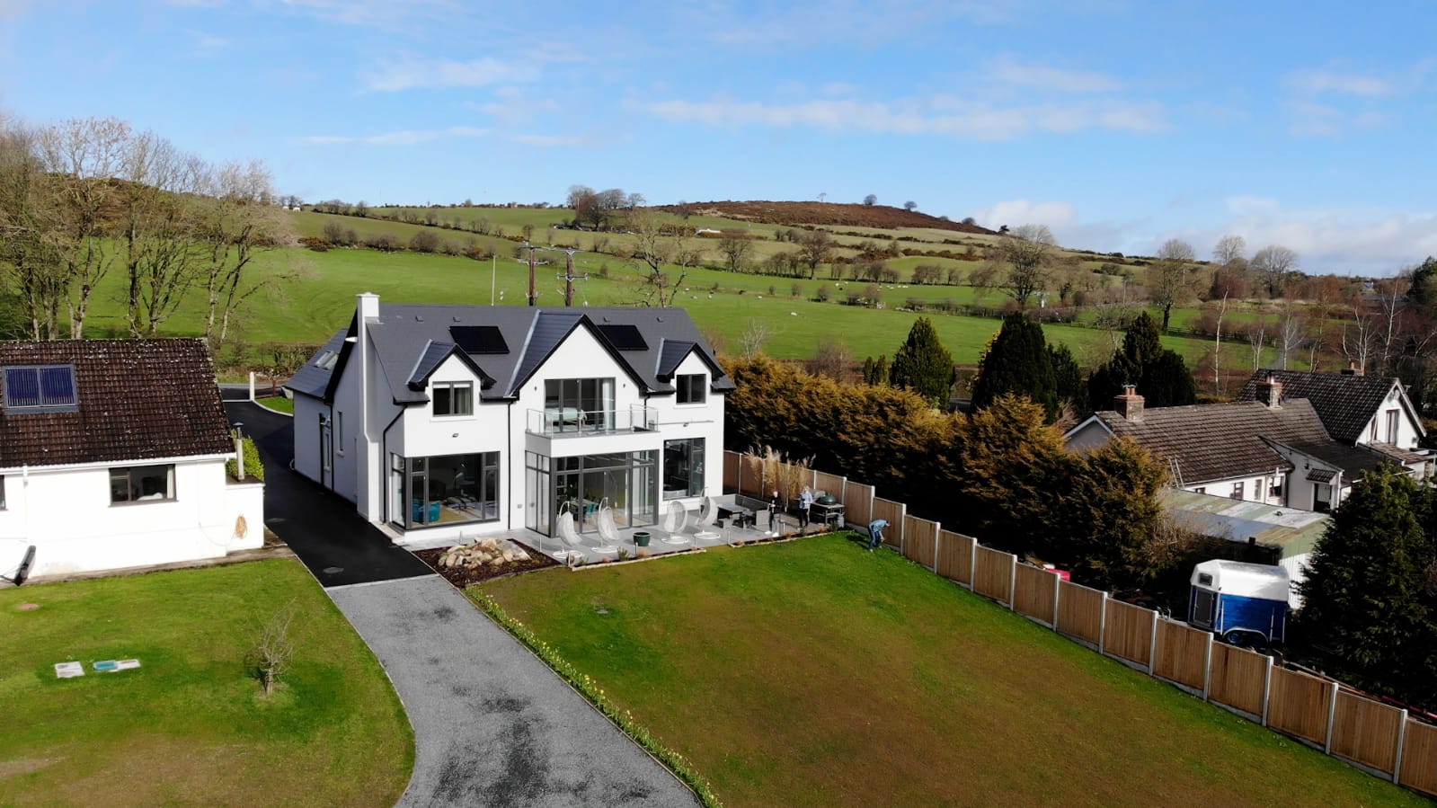 A white rendered house with dormer windows in the roof, a first floor balcony and full height glazing onto a patio, with green fields rising behind it
