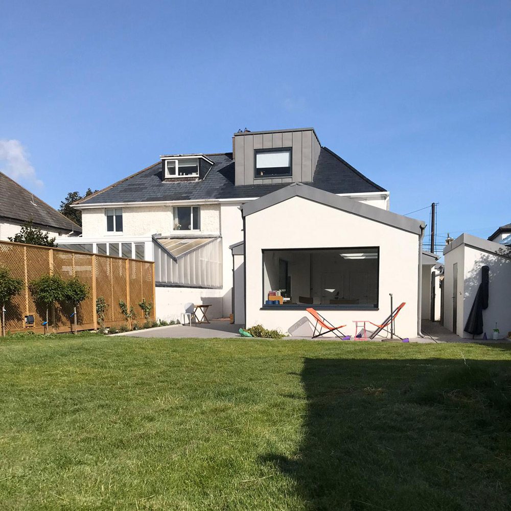 A rendered two storey house with a new dormer in the roof and a dark clad extension to the rear, seen across the back lawn
